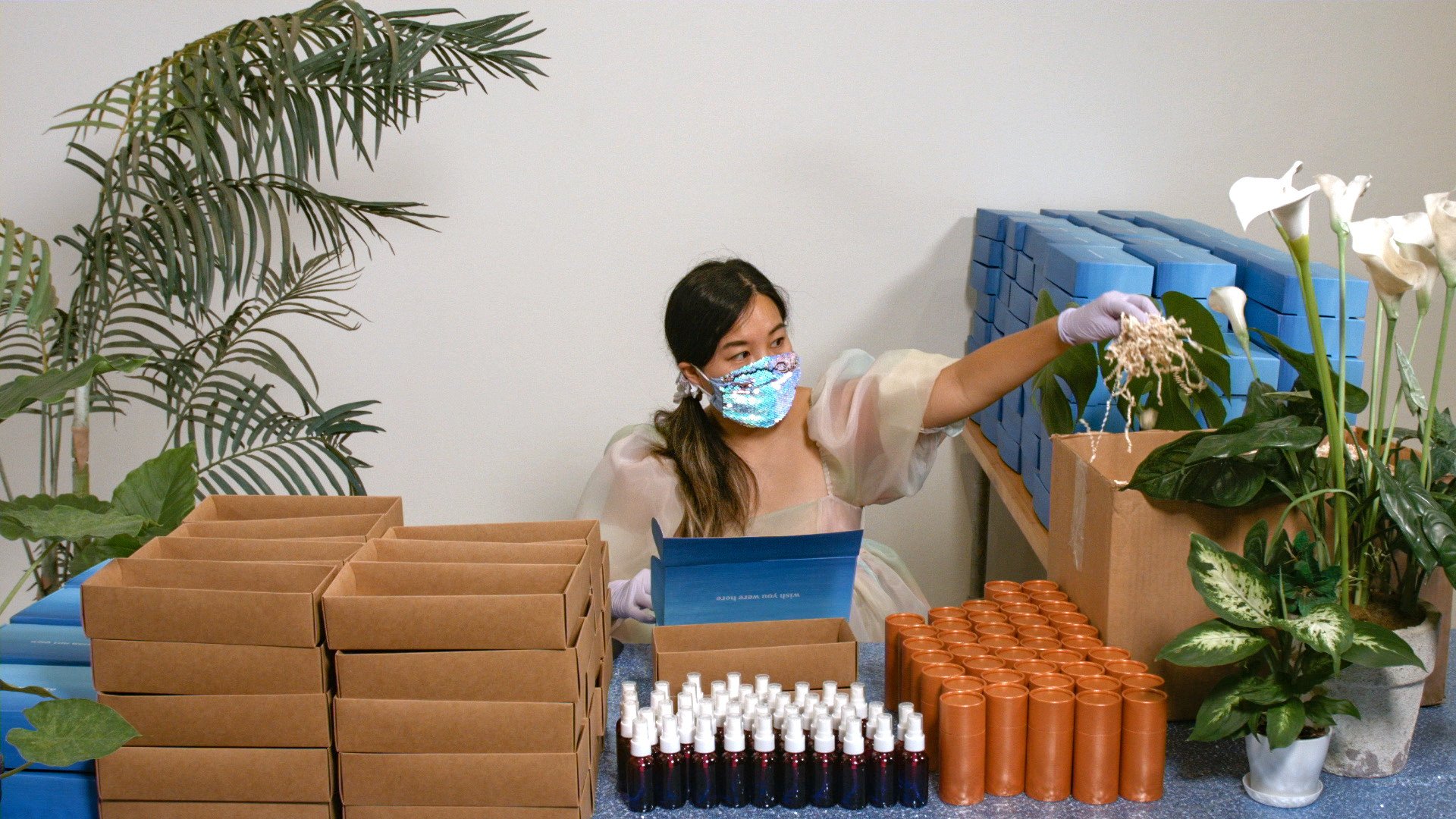 Woman wearing gloves and a sequined face mask packing objects at a table surrounded by cardboard boxes, bottles, plants, and stacks of blue packages.