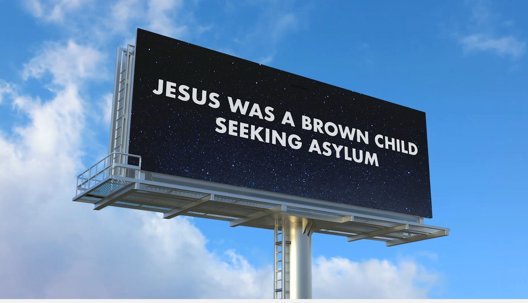 Roadside billboard against a blue sky reading “JESUS WAS A BROWN CHILD SEEKING ASYLUM” in white letters on a black starry background.