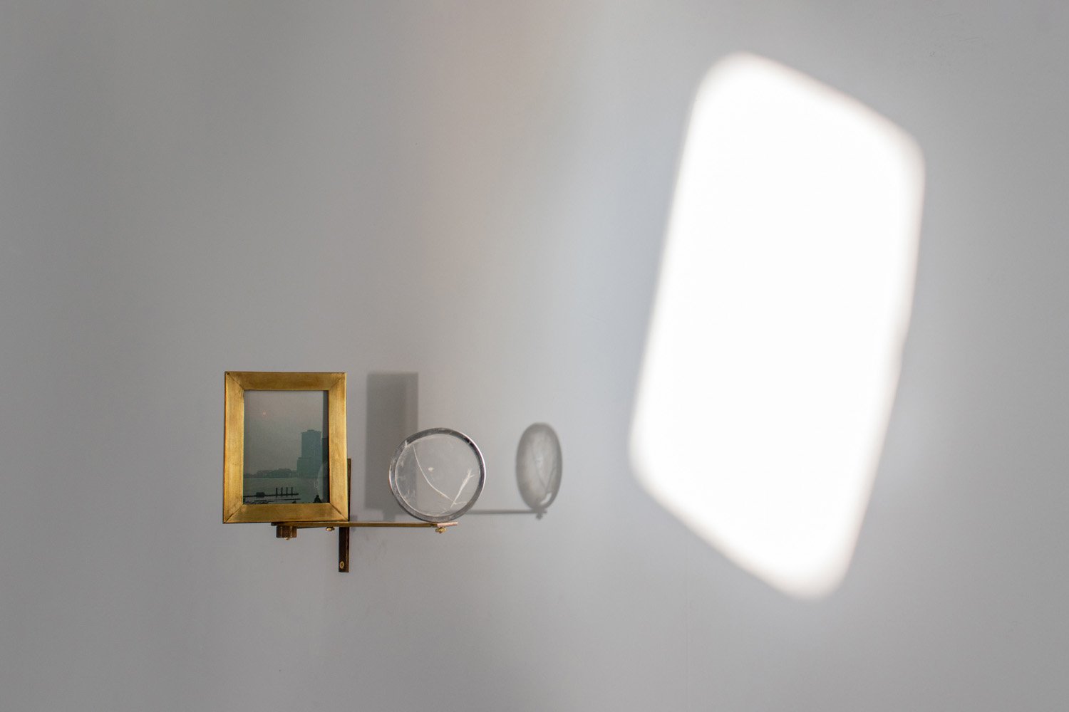 Installation view of a brass wall-mounted apparatus with a small framed window and circular lens casting a bright rectangle of sunlight onto a white wall.