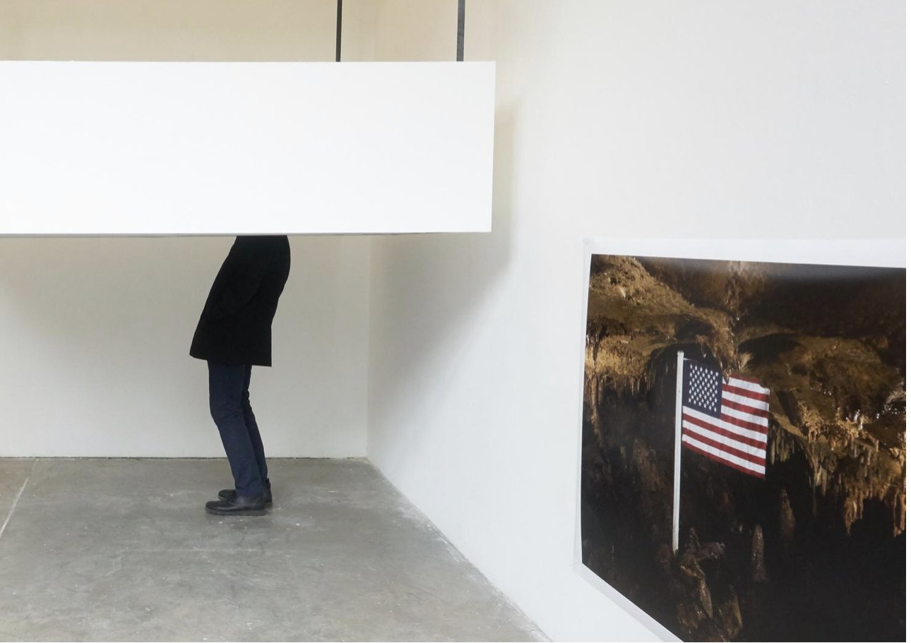 Installation view with a person partly obscured beneath a low white architectural structure and a photograph of an American flag inside a cave on the adjacent wall.