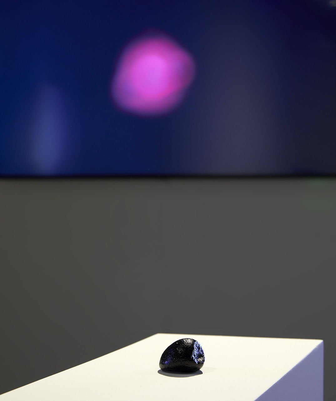 Installation view with a small dark iridescent sculpture on a white pedestal and a soft magenta form projected onto a dark wall behind it.