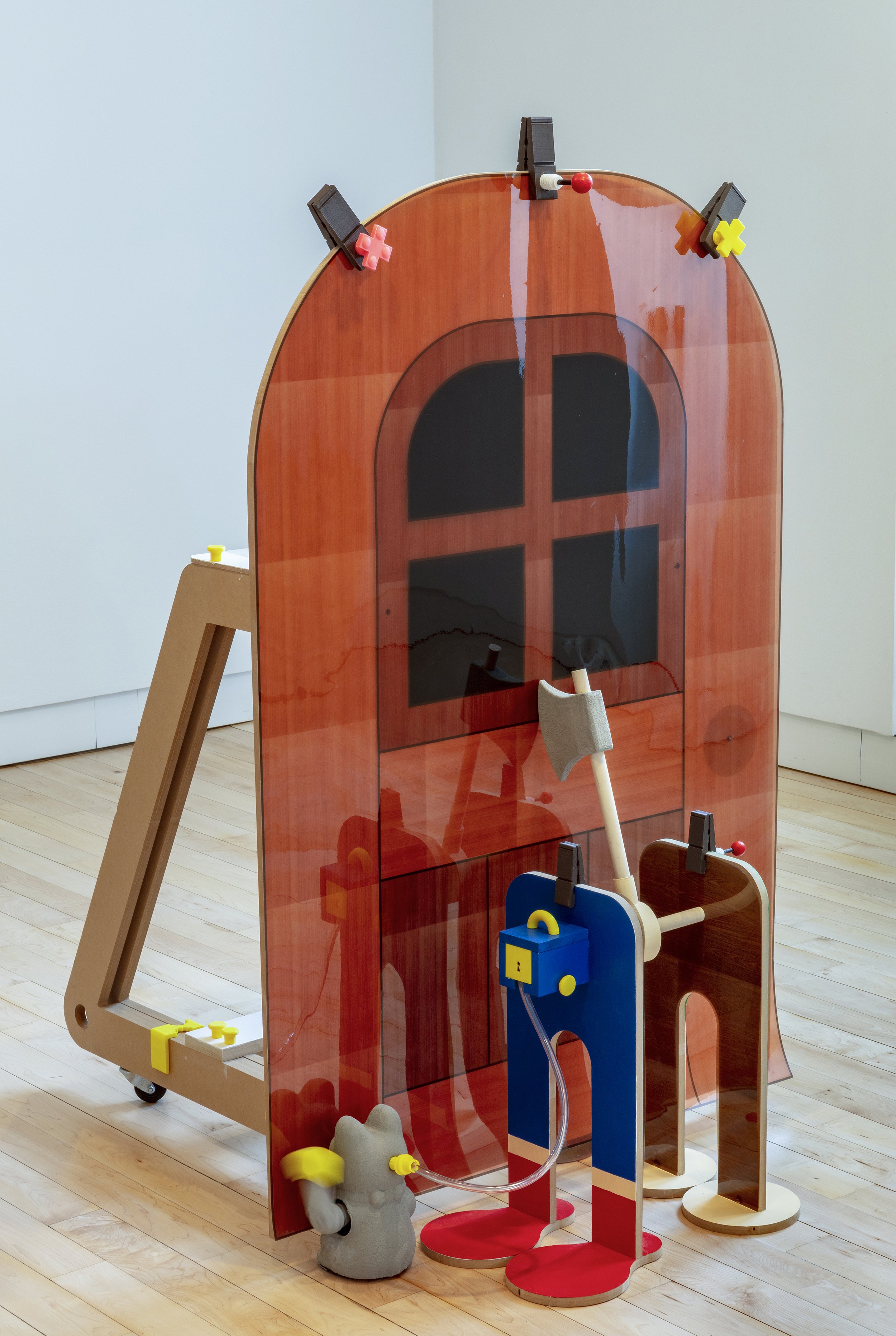 Toy-like sculpture built around a tall translucent orange house-shaped panel, with colorful clamps, a blue figure-like mechanism, tubing, wheels, and small molded components.
