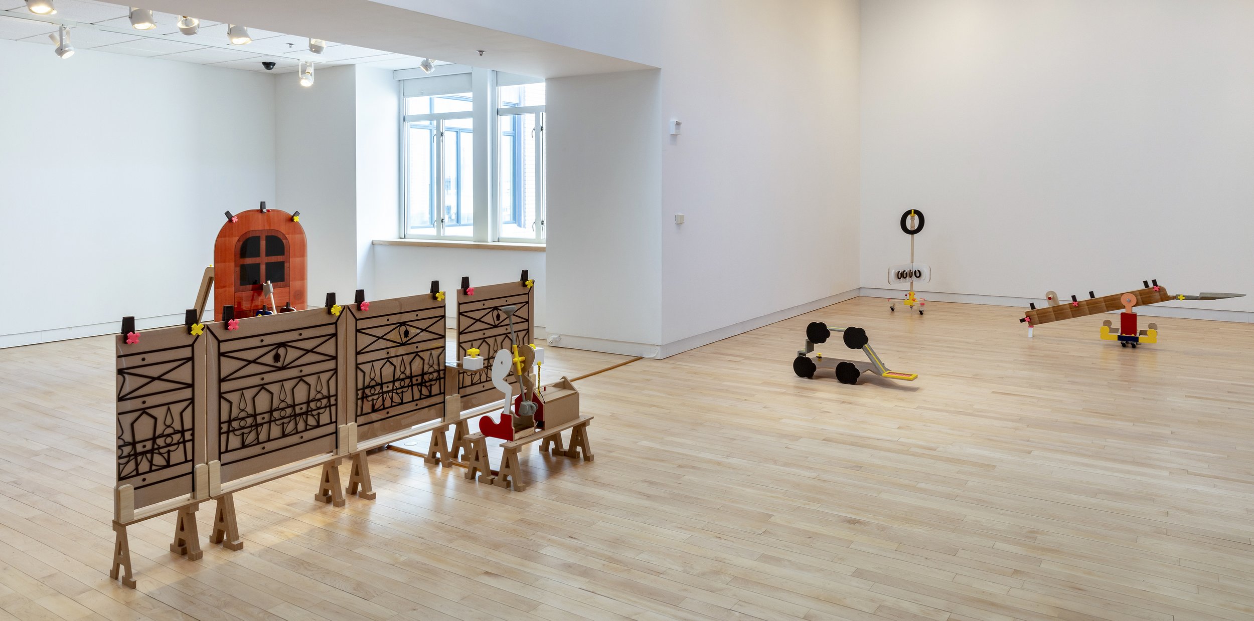Wide installation view of a gallery filled with toy-like sculptural objects, including cutout brown panels, benches, wheeled forms, and colorful mechanical structures.
