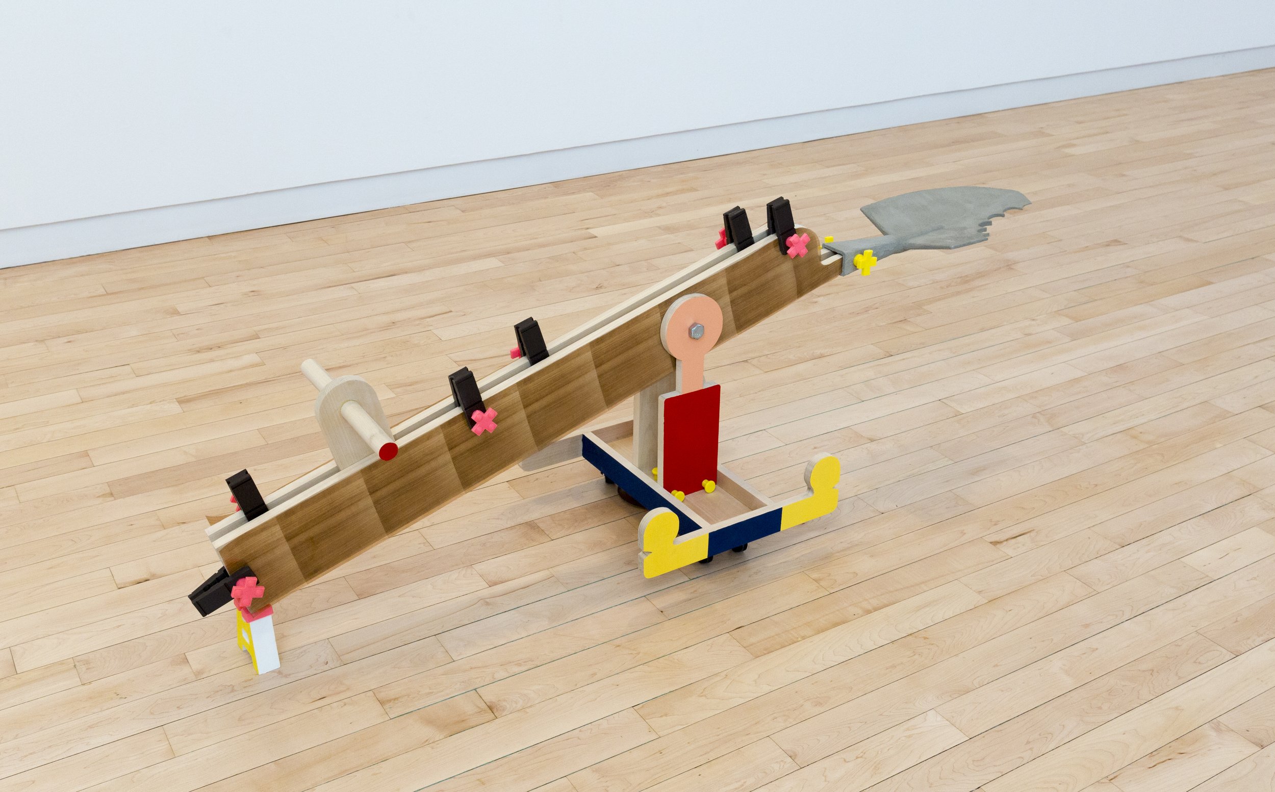 Long seesaw-like wooden sculpture balanced on a colorful wheeled base, with small clamps along the beam and a gray shovel-shaped form at one end.