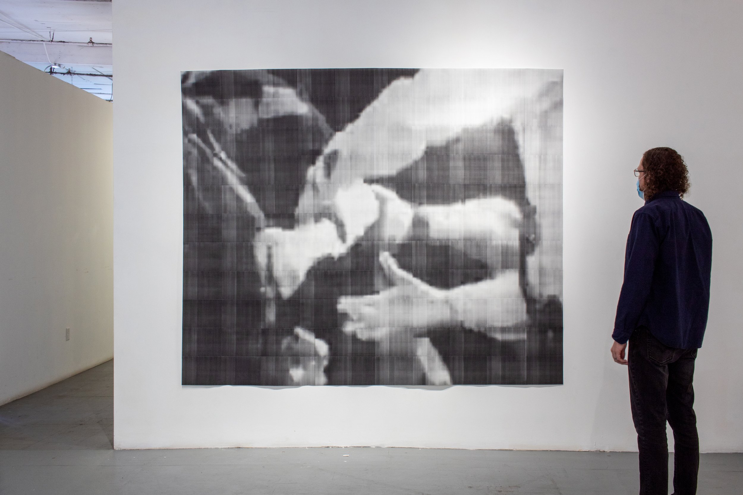Large black-and-white toner print showing a close-up of several arms and hands during a physical confrontation, viewed by a person standing beside the work.
