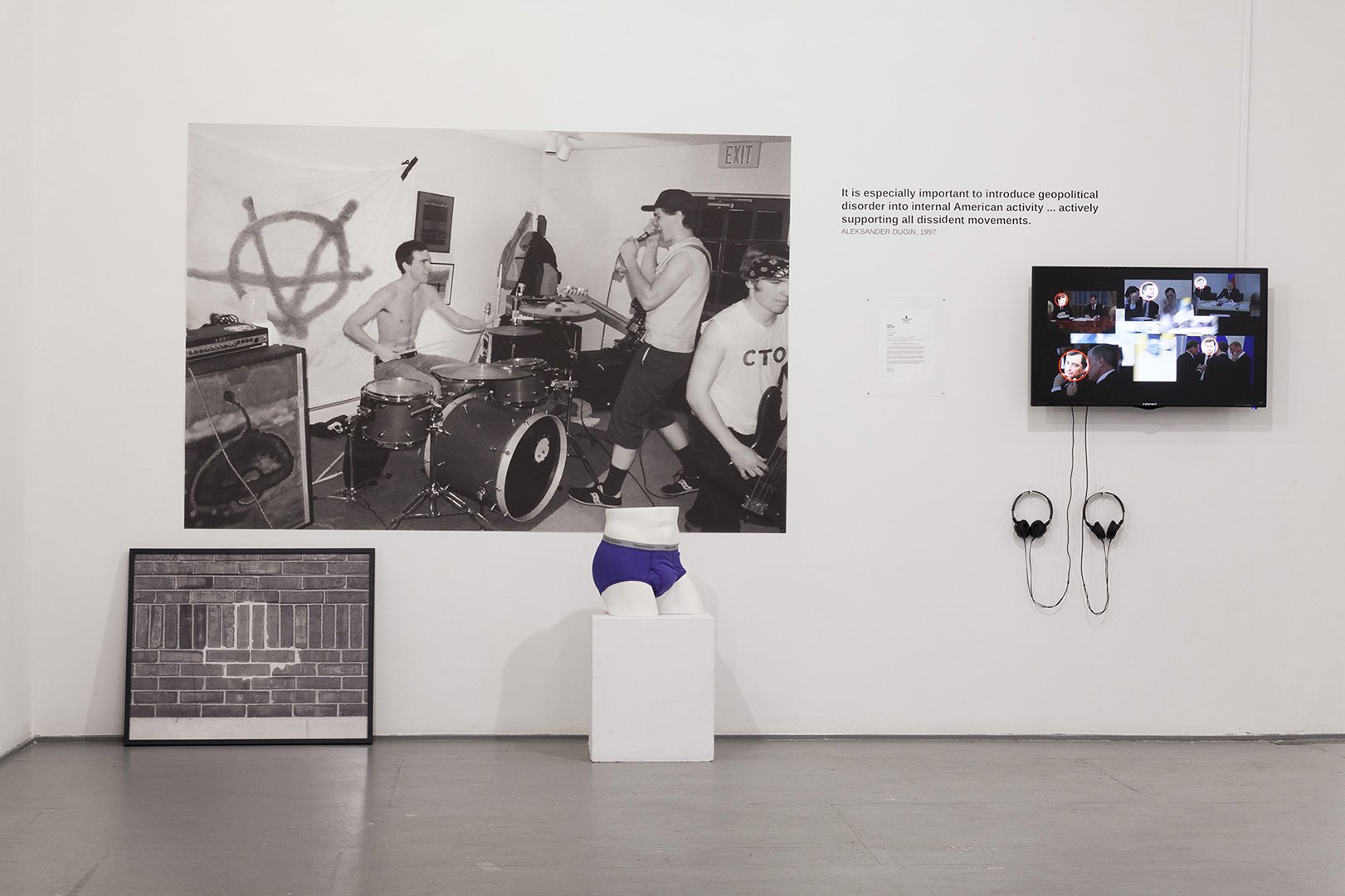 Installation view with a large black-and-white photograph of a punk band, a framed brick image, a sculpture of purple underwear on a pedestal, and a video monitor with two sets of headphones.