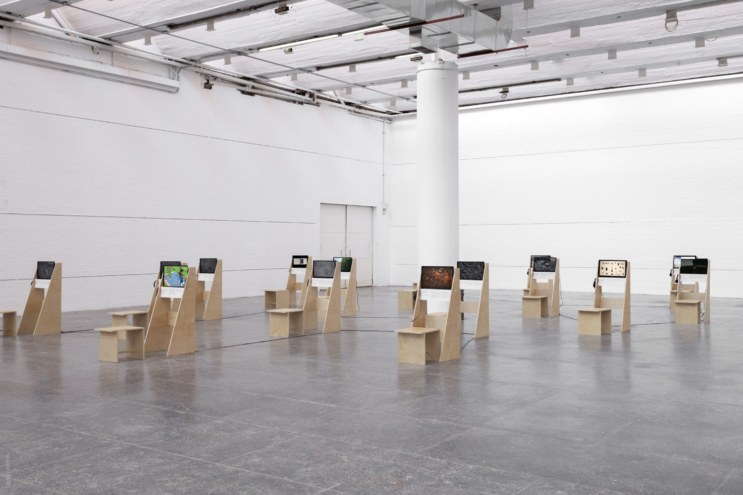 Installation view of Projected Ecologies at MUCA, showing rows of plywood video stations with monitors and stools in a white gallery.