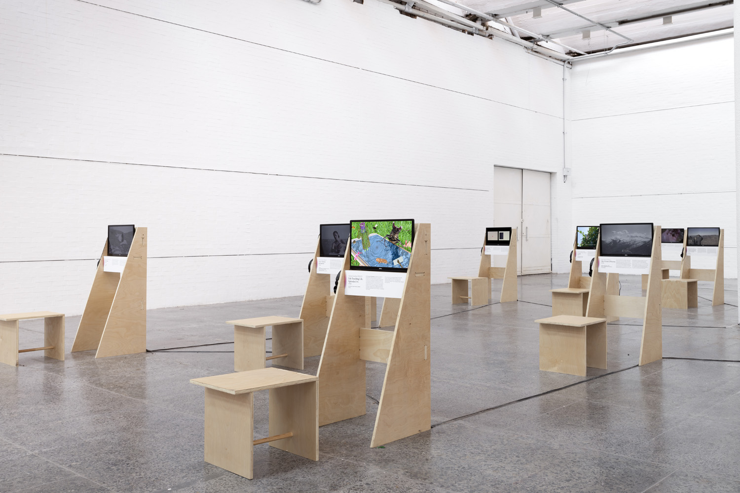 Installation view with a central video station showing a colorful map-like image, surrounded by additional monitors and stools.
