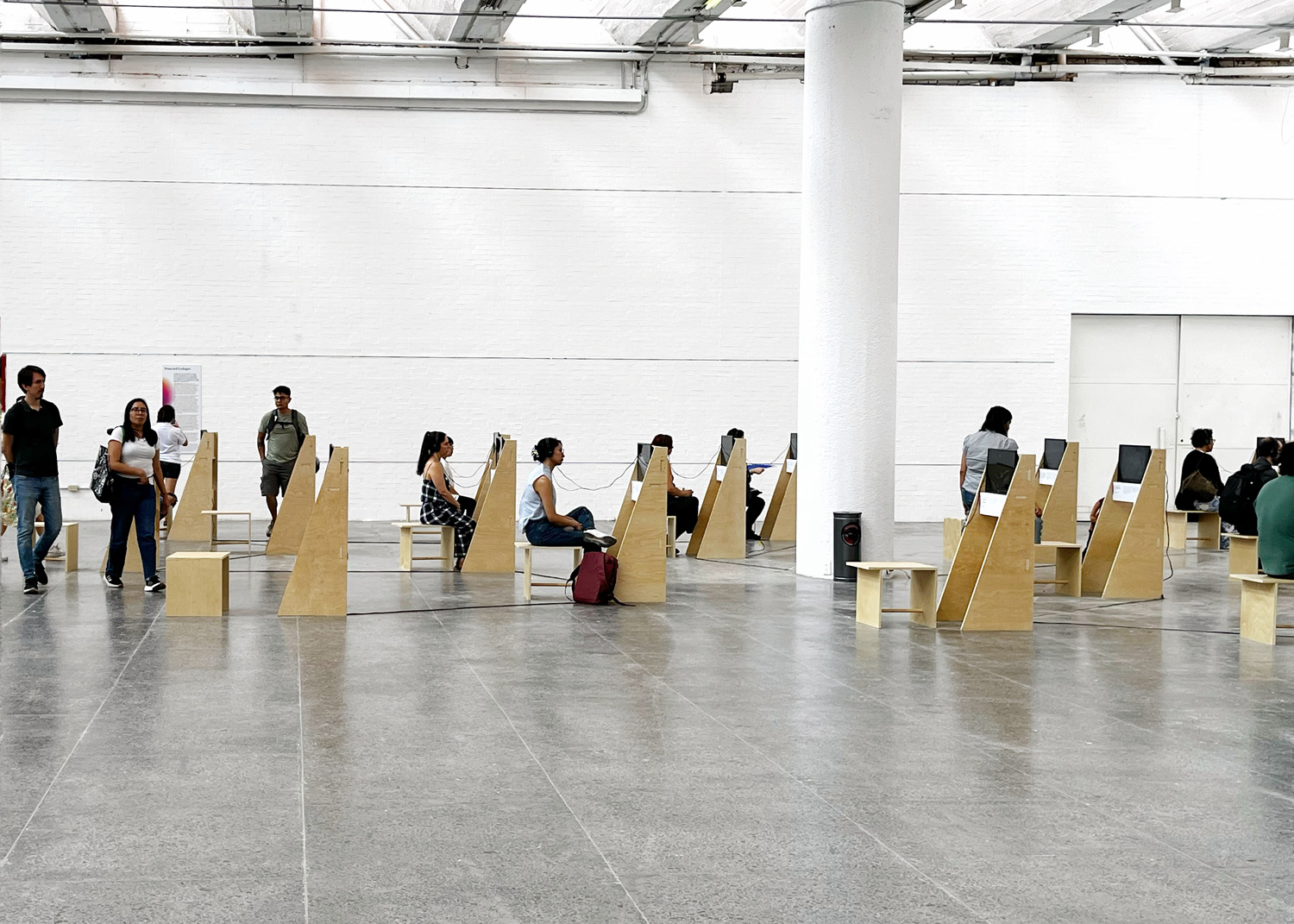 Side view of visitors seated and standing among rows of plywood video stations in Projected Ecologies.