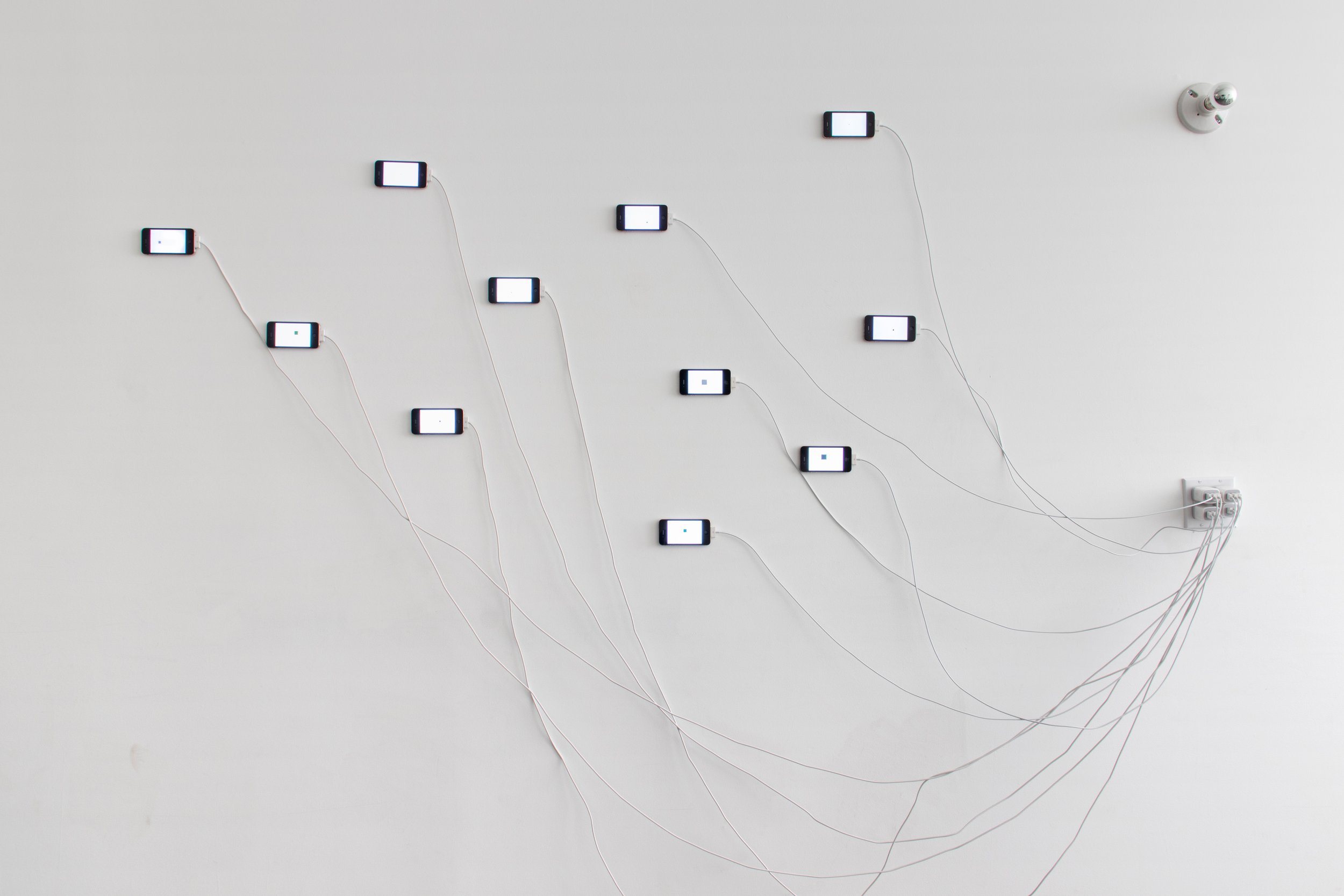 Wide installation view of eleven smartphones distributed across a white wall, connected by long charging cables that converge at an electrical outlet.