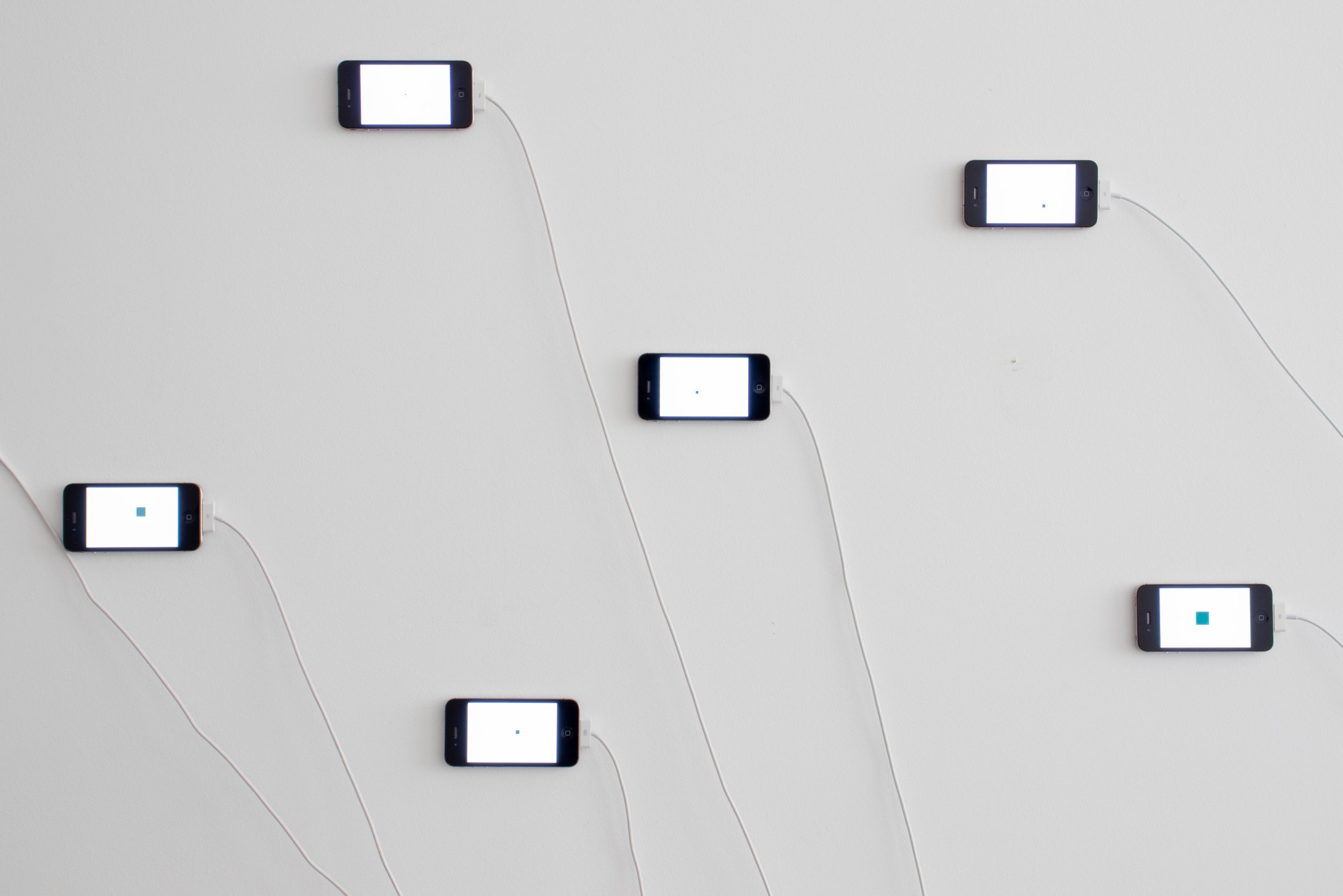 Installation detail showing several smartphones mounted horizontally on a white wall, each displaying a tiny colored square, with charging cords trailing downward.