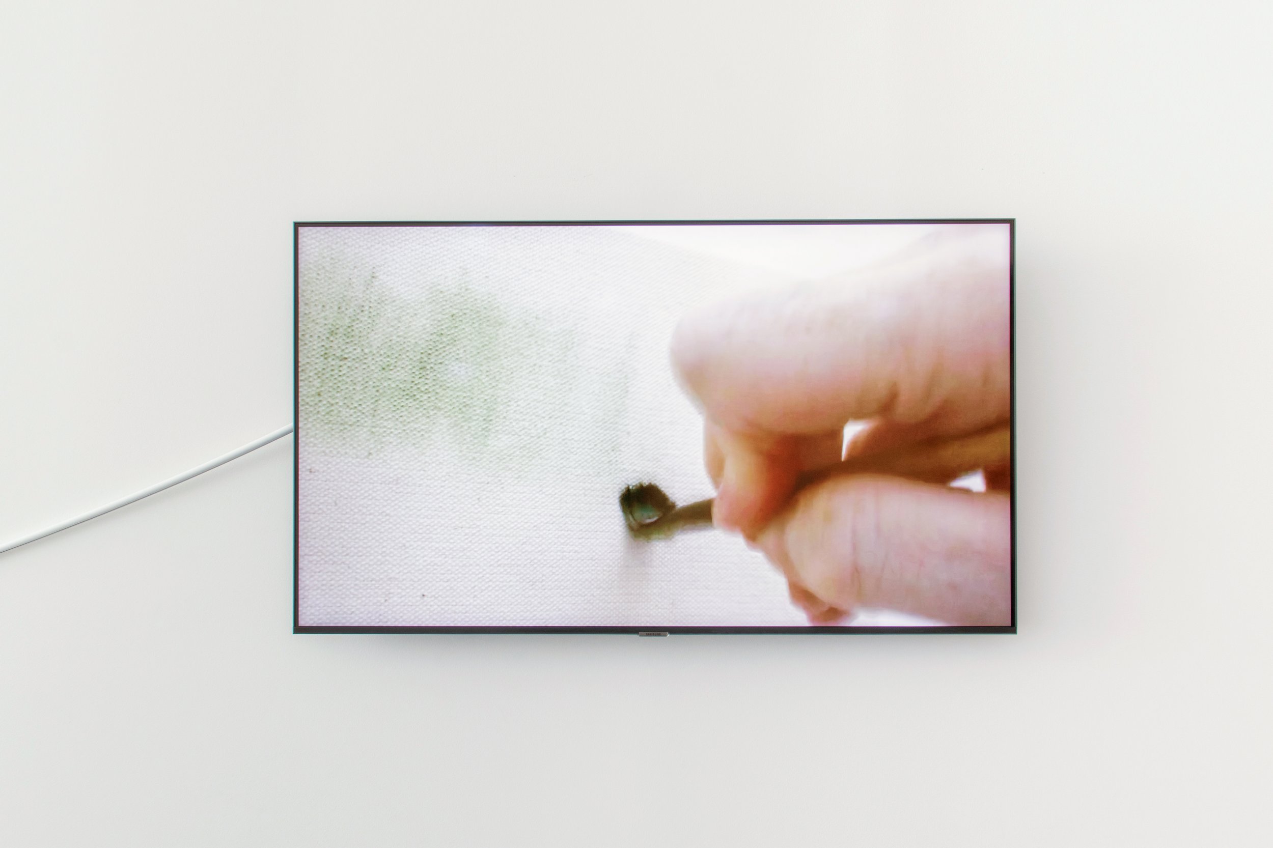 Video installation showing a close-up of a hand using a small tool to make a mark on the surface of a pale canvas.